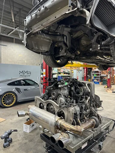 Porsche flat-six engine removed from the car on a stand under a raised 911